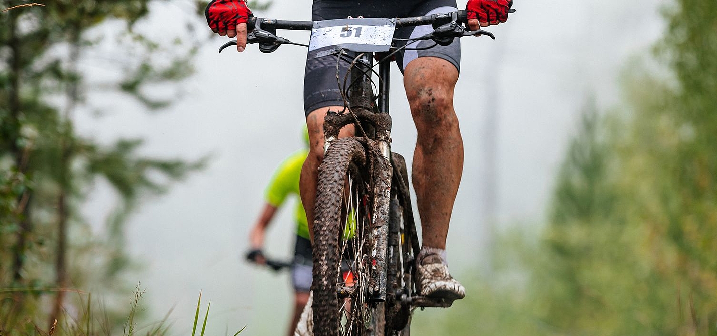 Header Mtb-Marathon Ein Mountainbike-Fahrer, verschmutzt und mit Startnummer am Lenker, sichtbar bis knapp oberhalb des Lenkers, fährt bergauf. Im Hinergrund ist ein weiterer Fahrer zu sehen.