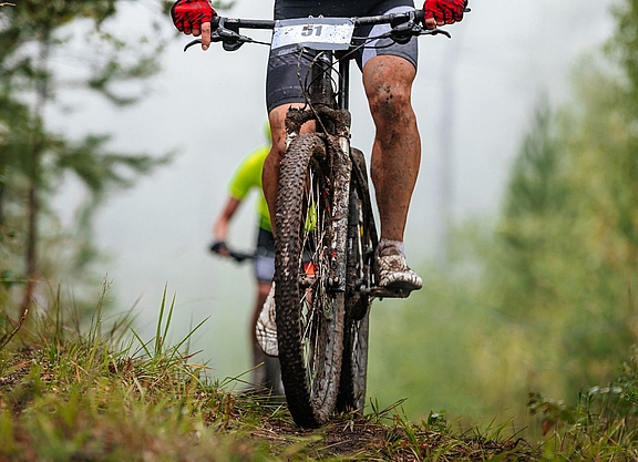 Header Mtb-Marathon Ein Mountainbike-Fahrer, verschmutzt und mit Startnummer am Lenker, sichtbar bis knapp oberhalb des Lenkers, fährt bergauf. Im Hinergrund ist ein weiterer Fahrer zu sehen.