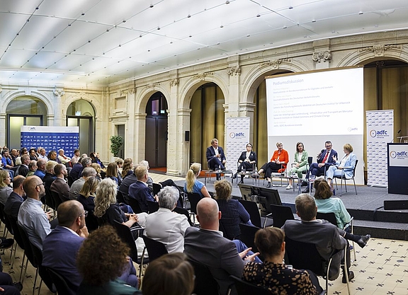 Fünf personen sitzen in offenem Halbkreis auf einer Bühne und diskutieren. Im Vordergrung Publikum auf Stühlen. 