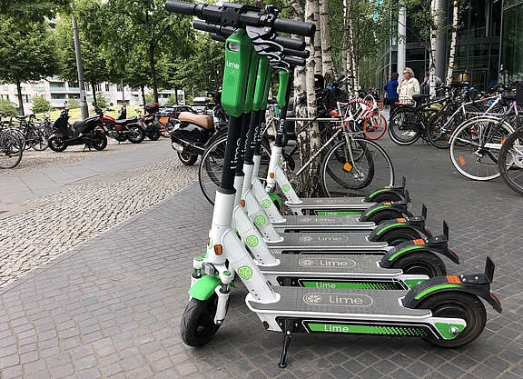 Im Vordergrund mehrere E-Scooter eines Verleihs, im Hintergrund mehrere Fahrräder, einige Personen und Gebäude