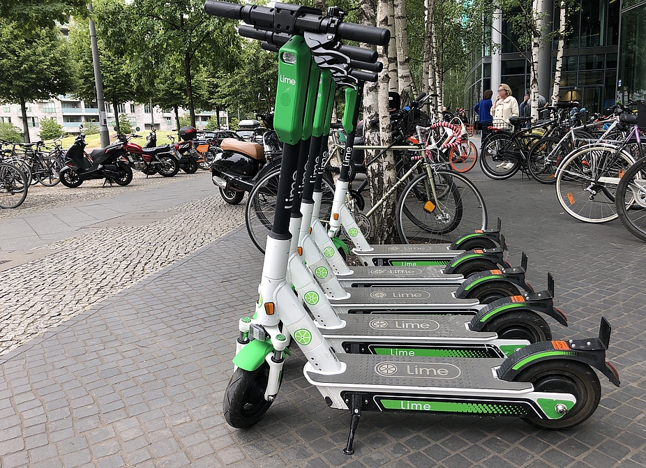 E-Scooter zum Ausleihen in Berlin Im Vordergrund mehrere E-Scooter eines Verleihs, im Hintergrund mehrere Fahrräder, einige Personen und Gebäude