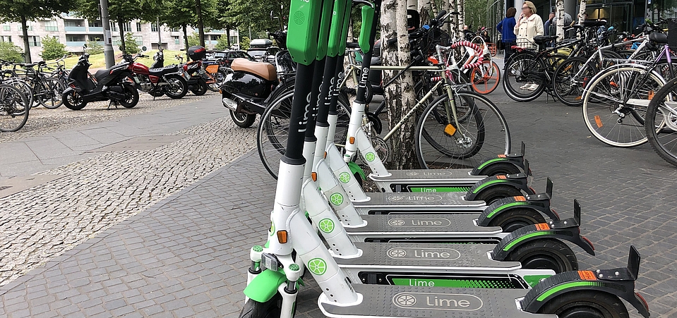 E-Scooter zum Ausleihen in Berlin Im Vordergrund mehrere E-Scooter eines Verleihs, im Hintergrund mehrere Fahrräder, einige Personen und Gebäude