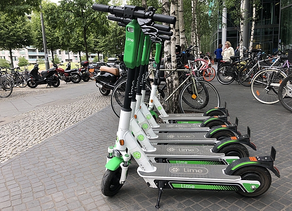 Im Vordergrund mehrere E-Scooter eines Verleihs, im Hintergrund mehrere Fahrräder, einige Personen und Gebäude