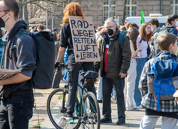 Demonstration im Freien. Im Vordergrund steht eine Person mit einem Fahrrad, die ein Schild mit der Aufschrift „Radwege retten Luft und Leben trägt“