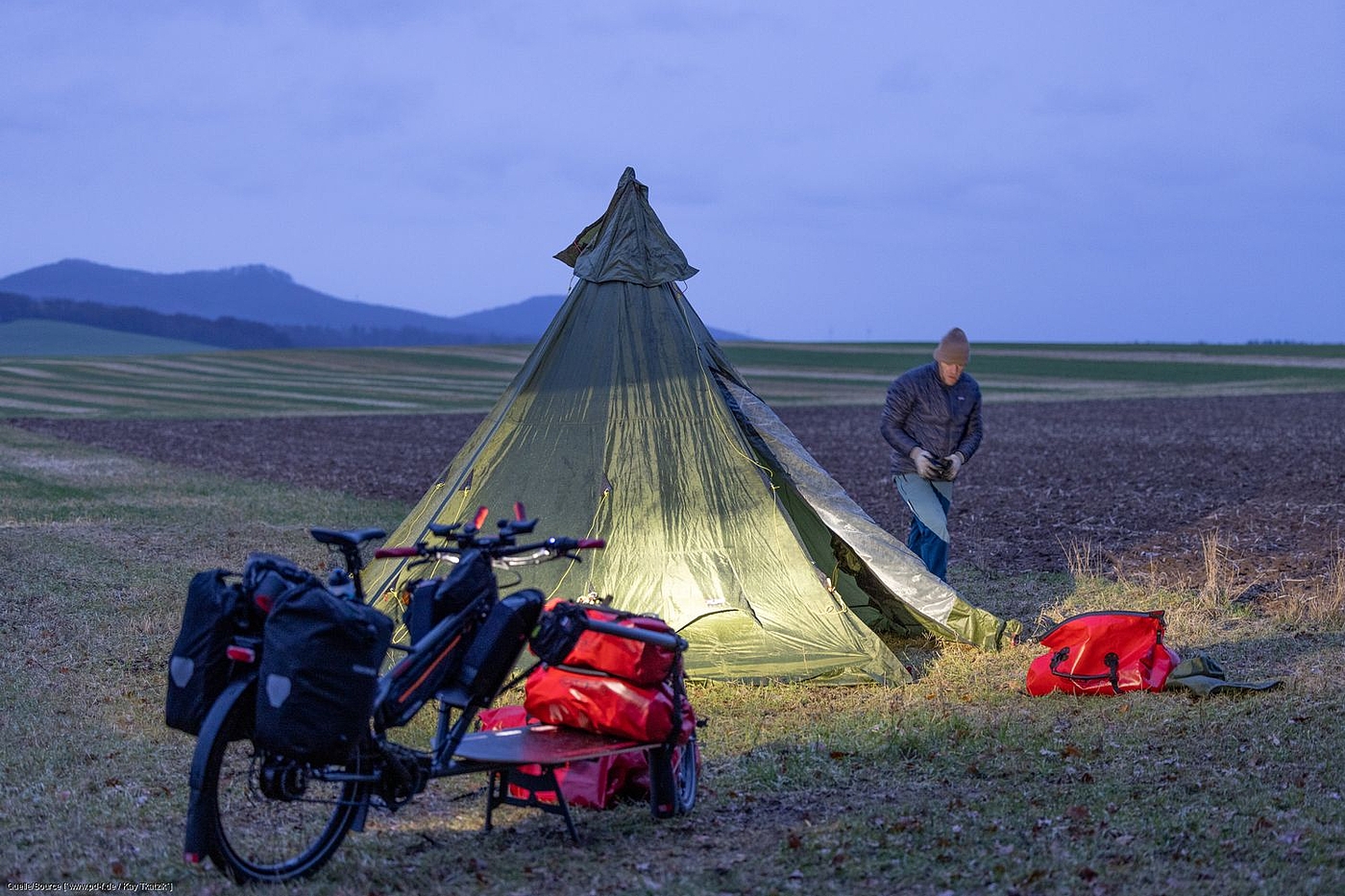 Workpacking: Übernachten im Zelt Ein Zelt steht auf einem Feld, davor ein Lastenrad. Gunnar Fehlau läuft um das Zelt herum, um Heringe zu befestigen.